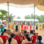 Young girls in bright dresses perform a line dance under a patterned canopy while adults and classmates watch from red chairs nearby